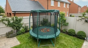 A photorealistic view of a 6ft trampoline in a small garden, neatly protected by a tailored dark green winter weather cover, with scattered autumn leaves and water droplets on the surface.