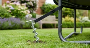 Illustration demonstrating how to secure an 8ft trampoline frame to lawn turf using heavy-duty ground anchor pegs and straps to withstand high winds.