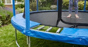 A close-up shot of an 8ft trampoline showing a child's legs inside the zip-up safety enclosure net, thick blue foam padding on the poles, and exposed high-tension steel springs.