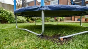 A close-up photograph focusing on the lower frame and legs of a blue and metal toddler trampoline on a green lawn. A small section of dark soil next to the metal leg reveals a distinct British 1p coin lying flat on the ground.