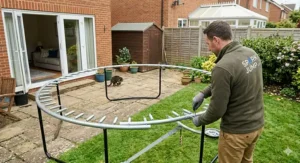 A man wearing an olive-green fleece assembling the large metal frame ring of an 8 foot trampoline in a typical British garden layout with a brick house and conservatory in the background.