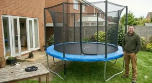 A man standing proudly next to a fully assembled 8 foot trampoline with a tall safety net enclosure in a typical British back garden with a brick home and fence line.